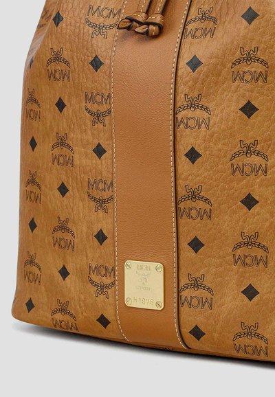 Sac MCM en cuir camel avec motif de logo noir, bande centrale cousue, plaque de marque dorée et détail de fermeture à cordon.