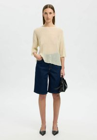 Cremfarbener, gerippter Strickoberteil mit drei Viertelärmeln, kombiniert mit dunkelblauen Denim-Bermudashorts. Das Model hält eine kleine schwarze Handtasche.