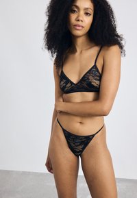 Zwart kanten lingerie set bestaande uit een bralette met dunne bandjes en een driehoekige uitsnede, gecombineerd met een bijpassende thong, beide met een bloemenpatroon.