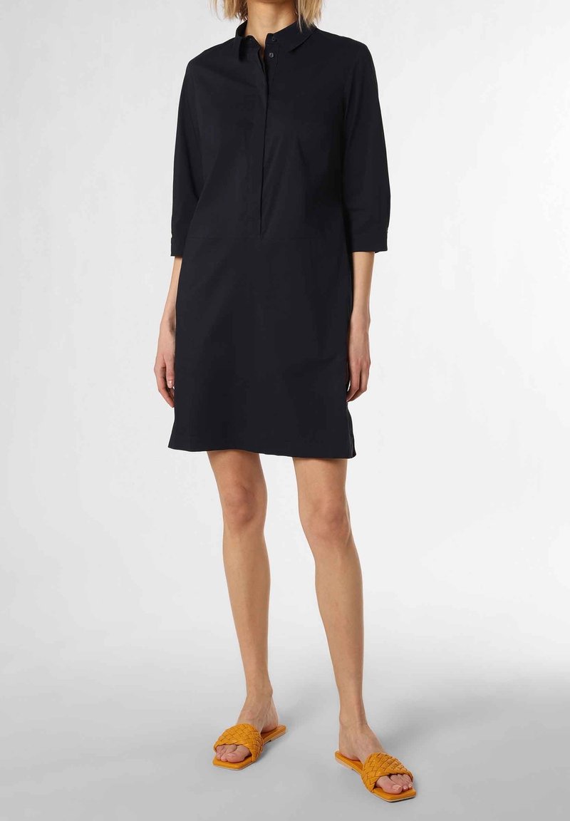 Kvinna som bär en knälång svart skjortklänning med trekvartslånga ärmar och orange vävda öppna tofflor som står på ett vitt golv.