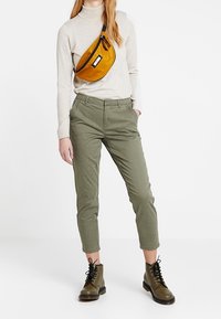 Oliwkowe, zwężające się spodnie z dekoracyjnym szwem po bokach, w połączeniu z jasnobeżowym golfem i żółtą torebką na talii. Brązowe buty sznurowane.