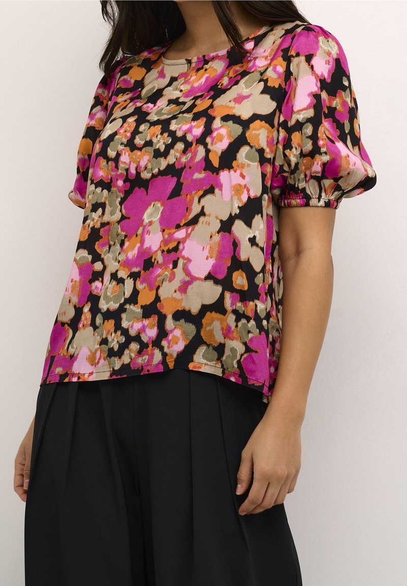 Femme portant un chemisier à manches courtes avec un motif floral lumineux en rose, orange, beige et noir, associé à un pantalon plissé noir.