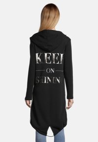 Sudadera larga con capucha negra que presenta la frase "KEEP ON SHINING" en impresión metálica plata en la parte posterior. Tejido suave con un detalle de dobladillo curvado.