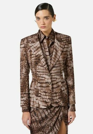 Donna che indossa un blazer marrone con motivo a fantasia e una gonna abbinata con spacco frontale, abbinati a orecchini dorati e capelli tirati indietro.