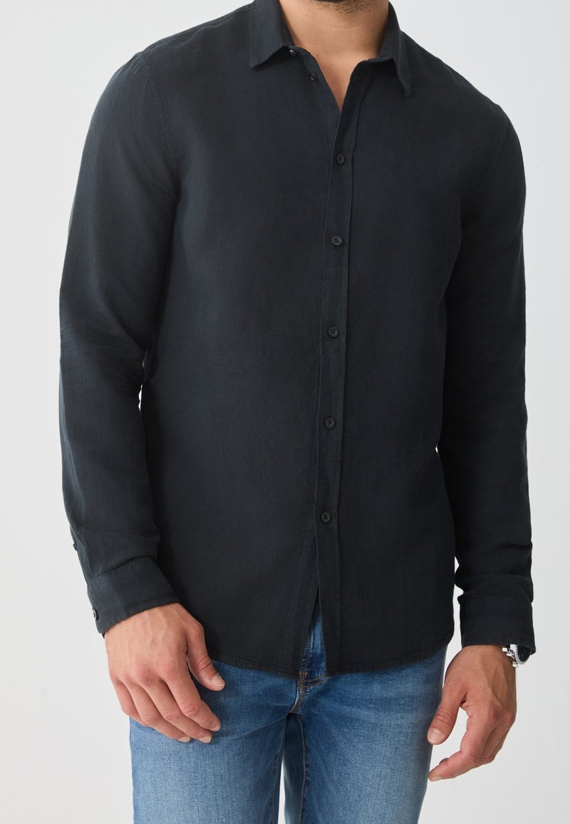 Homme portant une chemise noire à manches longues boutonnée et un jean bleu, debout devant un fond uni.