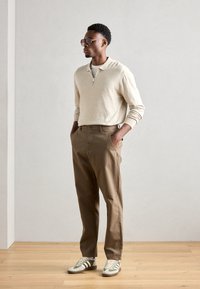 Svetilka v svetlo bež barvi z zadrgo, rjave chinos hlače in zeleni/beli športni čevlji. Slim fit, gladka tekstura in subtilne oblikovne podrobnosti.
