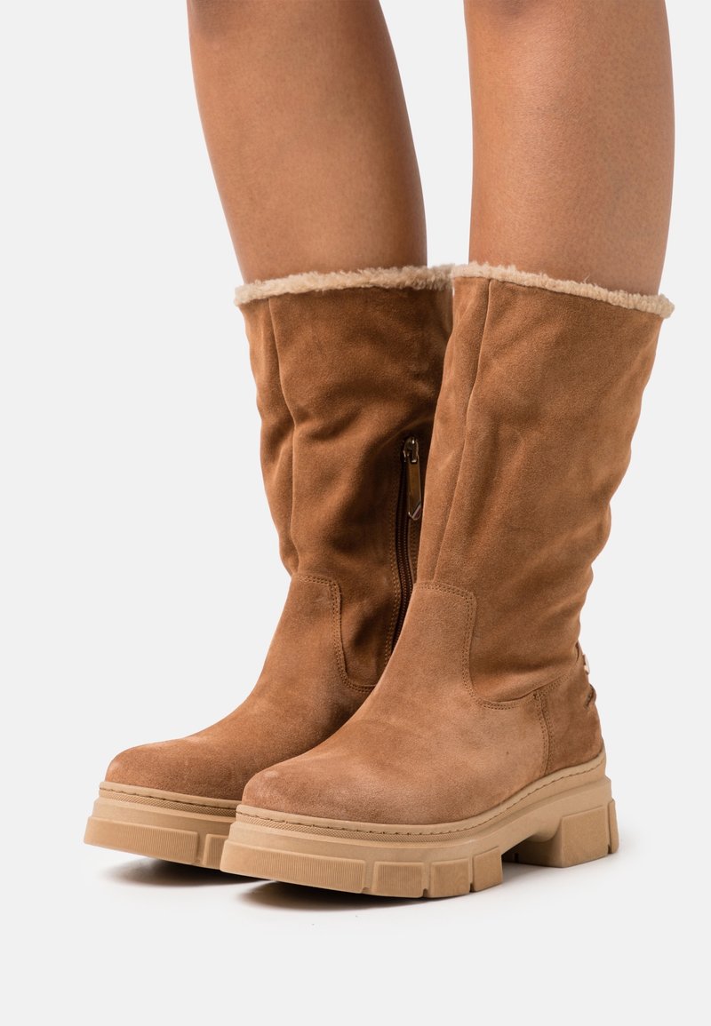 Bottes en daim marron montantes avec une doublure en fourrure, présentant une fermeture éclair sur le côté et une semelle épaisse en caoutchouc beige. Texture lisse, design décontracté.