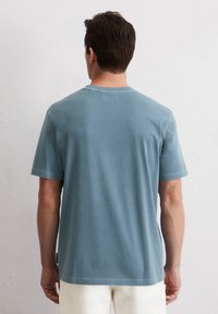 Kurzarm-T-Shirt aus weichem blauen Stoff, mit Rundhalsausschnitt, das in einem schlichten Schnitt gehalten ist und ein kleines Etikett an der linken Seitennaht aufweist.
