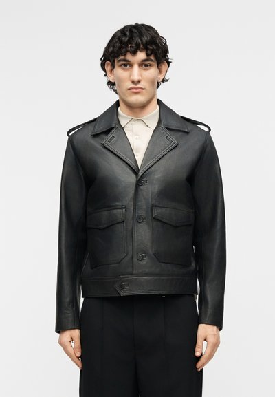 Νέος άνδρας με μαύρα σγουρά μαλλιά φοράει μαύρο δερμάτινο σακάκι με κουμπιά πάνω από μπεζ πουκάμισο με γιακά και μαύρο παντελόνι, κοιτάζοντας μπροστά, με απλό φόντο.