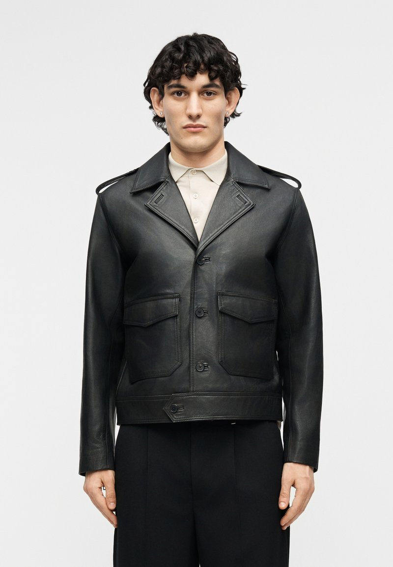 Young man with curled black hair wears black leather buttoned jacket over beige collared shirt and black pants, facing forward against plain background.