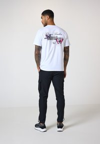 Weißes Baumwoll-T-Shirt mit einem Grafikdruck von bunten Schmetterlingen und Text auf der Rückseite. Kombiniert mit schwarzen Cargo-Hosen und Sportschuhen.