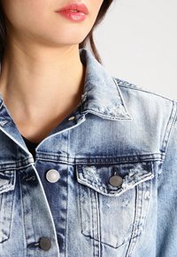 Jeansjacka i ljusblått med en slitstark textur, med metallknappdetaljer och bröstfickor. Närbild på kragen och tyget.