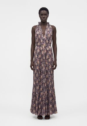 Γυναίκα που φοράει ένα αμάνικο, μακρύ μέχρι το πάτωμα φόρεμα με λουλούδια, βαθύ ντεκολτέ σε σχήμα V και λεπτή ζώνη, στέκεται με τα χέρια χαλαρά στο πλάι.