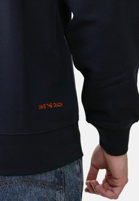 Felpa navy realizzata in misto cotone, con polsini a costine e un logo arancione "SAVE THE DUCK" visibile vicino all'orlo, vestibilità standard.