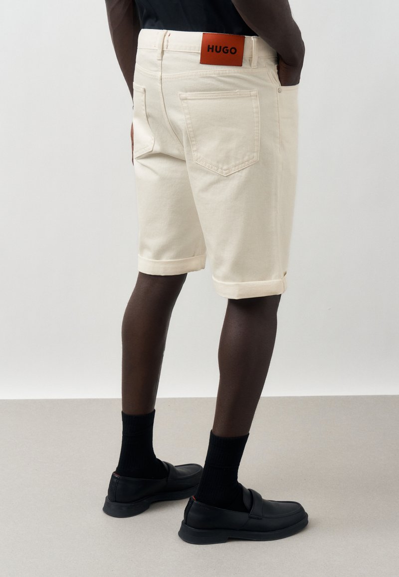 Άτομο που φοράει κρεμ σορτς με γυρισμένο τελείωμα και πίσω τσέπες, με πορτοκαλί ετικέτα "HUGO", συνδυασμένα με μαύρες κάλτσες και μαύρα λουστρίνια.