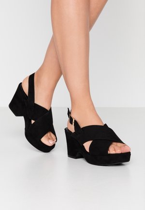 Czarne zamszowe sandały na koturnie z krzyżującymi się paskami, otwartą noskiem i regulowanym paskiem na kostkę z zapięciem w złotym kolorze.