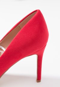Czerwone zamszowe buty na wysokim obcasie z ostrym noskiem i smukłym obcasem typu stiletto. Tekstura wydaje się gładka, a styl jest elegancki i nowoczesny.