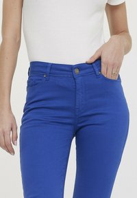 Jean en denim bleu avec taille mi-haute, design à cinq poches et quincaillerie dorée, présentant une texture lisse et une silhouette ajustée.