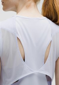 Close-up van een persoon in een witte mouwloze top met meshpanelen en een grote sleutelgatuitsparing op de bovenrug.