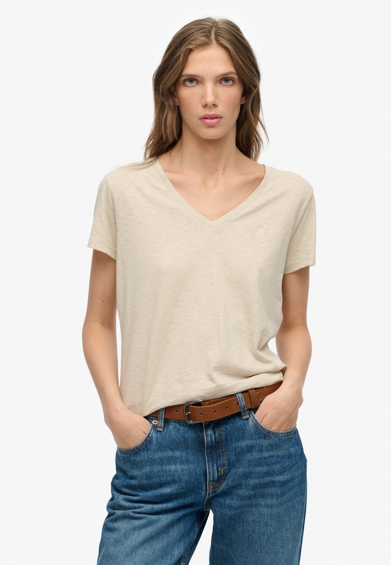 Beige V-hals t-shirt van zachte stof met korte mouwen, gecombineerd met blauwe denim jeans en een bruine leren riem. Minimale merkdetails aanwezig.