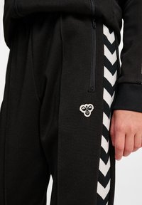 Zwarte joggerbroeken gemaakt van een zachte, gestructureerde stof, met een wit chevronpatroon langs de zijkanten en een klein geborduurd logootje.