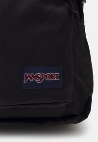 Čierny látkový ruksak s hladkou textúrou, sťahovacou šnúrkou a obdĺžnikovou nášivkou loga Jansport v námorníckej modrej a červenej farbe.