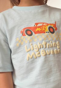 Šviesiai mėlynas trumpomis rankovėmis marškinėliai, kuriuose pavaizduota raudona lenktyninė mašina su ugnies akcentais ir tekstu "Lightning McQueen" kremine spalva.