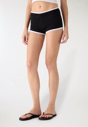 Pantaloncini neri elasticizzati con rifiniture bianche, dal design aderente e dalla texture liscia, che arrivano a metà coscia, abbinati a infradito nere.