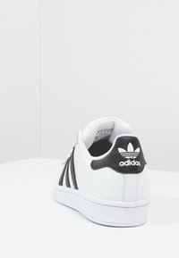Vita Adidas-sneaker med svarta ränder och logotyp på hälen, visad från bakre högra vinkel på vit yta och ljusgr å bakgrund.