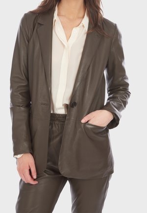 Persona che indossa un blazer in pelle marrone scuro e pantaloni abbinati con una camicia bianca abbottonata, una mano nella tasca del blazer.