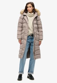 Manteau long en duvet beige clair avec capuche doublée de fourrure, texture matelassée, fermeture éclair sur le devant et fentes latérales. Associé à un jean bleu clair et des chaussures noires.