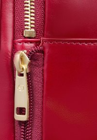 Rode leren ritstas met een goudkleurige rits en trekker. Heeft een gladde textuur en stiksel langs de rand.