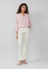 Chemisier rose clair à manches longues avec un décolleté en V, associé à un pantalon droit blanc et des sandales à talons avec lanières blanches. Textures lisses partout.