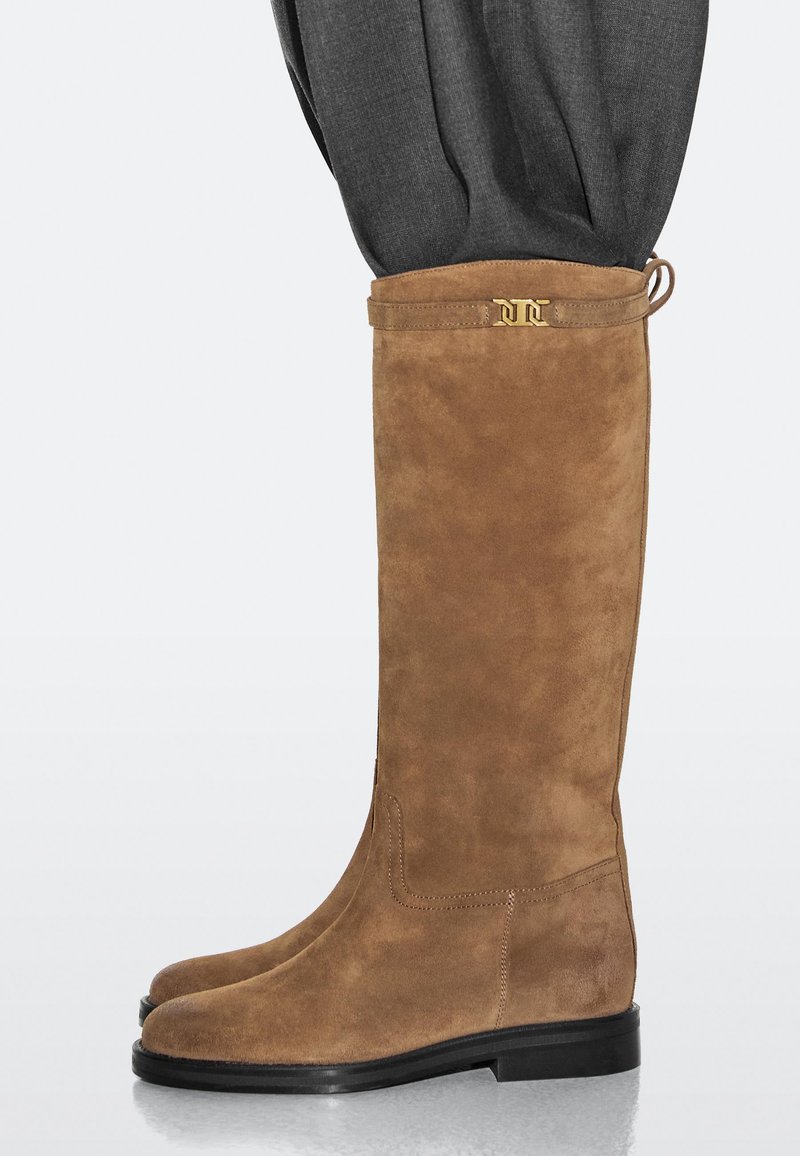 Wildleder-Braune kniehohe Stiefel mit abgerundeter Spitze, flacher schwarzer Sohle und einem goldenen Akzent am Schaft. Weiche Textur, minimale Nähte.