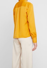 Žlutá hedvábná košile s manžetami na knoflíky, s plisovaným zadním dílem a hladkou texturou. Kombinována s krémovými kalhotami.