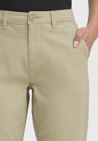 Beige kalhoty s hladkým povrchem, se zapínáním na jeden knoflík, předními kapsami a poutky na opasek, zobrazené v pase.