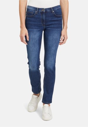 Vrouw die medium-wash slim-fit blauwe spijkerbroek draagt met voorzakken en witte sneakers, staand tegen een effen achtergrond.