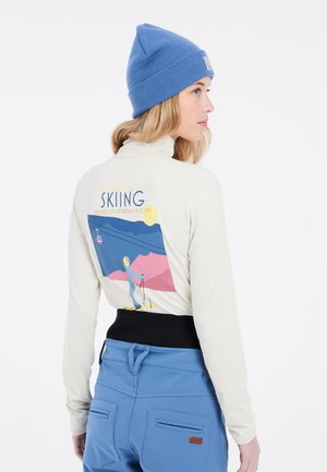 Langarmige crèmekleurige top met een ski-afbeelding op de achterkant, gecombineerd met blauwe skibroek met zakken en een zwarte tailleband.