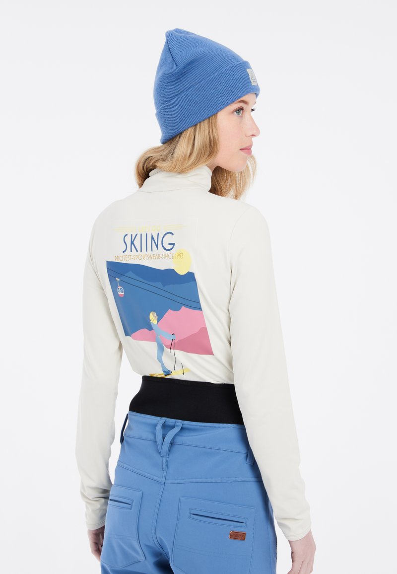 Langarmige crèmekleurige top met een ski-afbeelding op de achterkant, gecombineerd met blauwe skibroek met zakken en een zwarte tailleband.