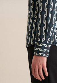 Camicia con motivo a catena in teal, bianco e blu scuro. Polsini con bottone e una texture di tessuto liscia. Pantaloni neri uniti visibili sotto.