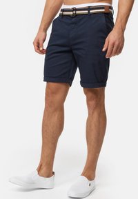 Marineblaue Baumwollshorts mit umgeschlagenen Säumen, ausgestattet mit einem mehrfarbigen gestreiften Stoffgürtel und einer metallischen Schnalle. Kombiniert mit weißen Slippern.
