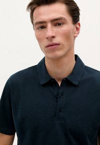 Námořnická modrá polokošile s límečkem a jezdci na tři knoflíky. Má hladkou texturu a krátké rukávy. Jednoduchý, čistý design.