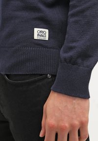Granatowy sweter z widocznymi rękawami i dołem oraz etykietą "ORIGINALS by Jack & Jones", w połączeniu z czarnymi spodniami i opadającą ręką.