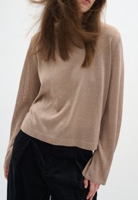 Femme portant un haut beige ample à manches longues et un pantalon noir, cheveux soufflés sur le visage, debout devant un fond clair uni.