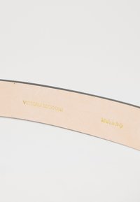 Lærinteriør i en lys beige belte med preget gulltekst: "Victoria Beckham" og "Made in Italy." Kantet i svart.