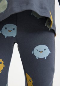 Mørkeblå leggings lavet af blødt stof, prydet med et gentaget tegneserie-mønster af smilende lyseblå figurer og gule former.