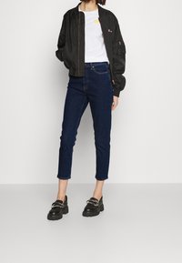 Svart bomberjacka, vit t-shirt med gult märke, mörkblå slim jeans och kraftiga svarta loafers med metalliska accenter.