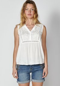Blusa bianca senza maniche con design ricamato sul davanti, caratterizzata da un scollo a V e un tessuto strutturato. Abbinata a pantaloni shorts in denim.