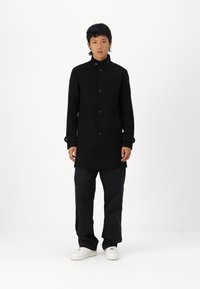 Abrigo negro largo con un cuello alto, cierre de botones en una sola fila, bolsillos laterales y acentos en los puños en contraste, combinado con pantalones negros holgados y zapatillas blancas.