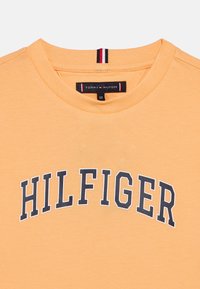 Πορτοκαλί βαμβακερό t-shirt με στρογγυλή λαιμόκοψη και το έντονο, ναυτικό μπλε λογότυπο "HILFIGER" στο στήθος. Περιλαμβάνει λεπτομέρειες ετικέτας της μάρκας.